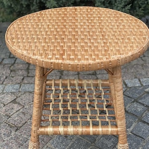May include: A round, woven wicker side table with a lower shelf. The table has a natural, light brown color and is made of woven wicker material. The table is approximately 20 inches in diameter and 20 inches tall.