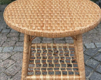 May include: A round, woven wicker side table with a lower shelf. The table has a natural, light brown colour and is made of woven wicker material. The table is approximately 50 cm in diameter and 50 cm tall.
