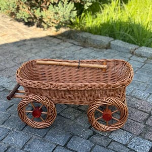 Op de afbeelding: Een bruine rieten wagen met een rechthoekige mand en twee spaakwielen. De wielen hebben rode naven. De wagen heeft een houten handvat en staat op een stenen ondergrond.