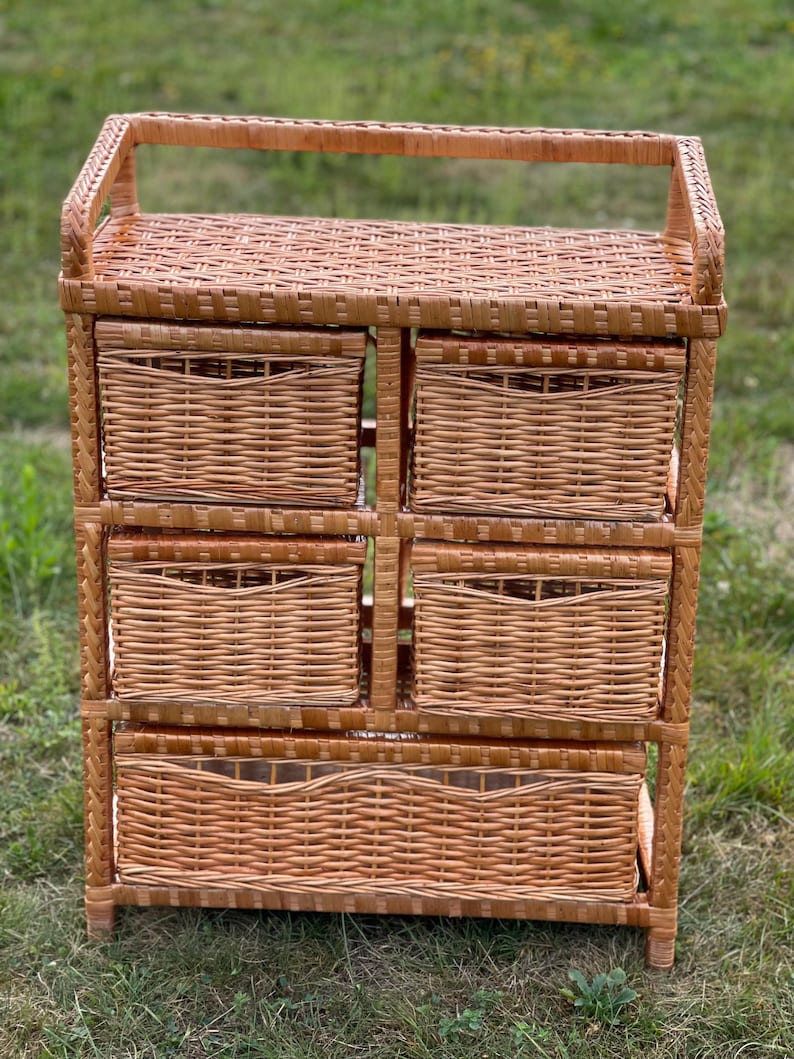 May include: A light brown wicker storage unit with six woven drawers and a top shelf. The unit has a handle on top and is made of woven wicker. The drawers are rectangular and have a woven pattern. The unit is standing on a grassy surface.