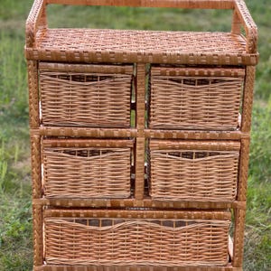 May include: A light brown wicker storage unit with five woven baskets. The unit has a rectangular shape with a handle on top. The baskets are of varying sizes, suitable for organizing items. The unit is made of natural materials.