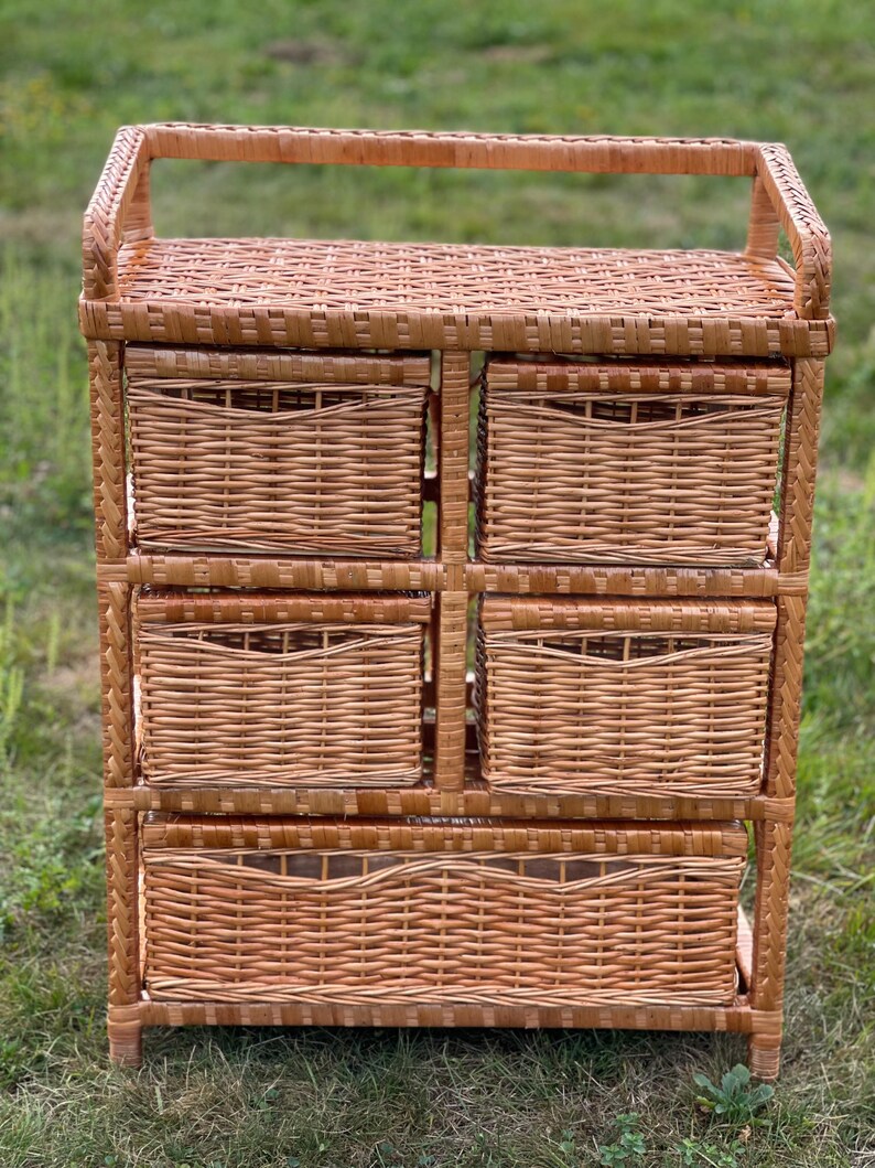 May include: A light brown wicker storage unit with five woven baskets. The unit has a rectangular shape with a handle on top. The baskets are of varying sizes, suitable for organizing items. The unit is made of natural materials.