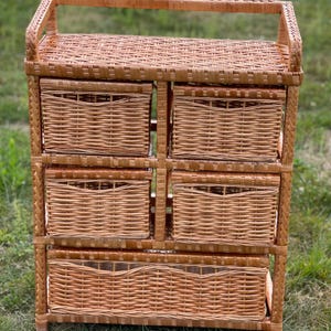 May include: A light brown wicker storage unit with six woven drawers and a top shelf. The unit has a handle on top and is made of natural wicker material. The drawers are rectangular and the unit is standing on a grassy surface.
