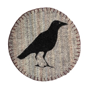 May include: A round, gray felt coaster with a black crow silhouette in the center. The crow is standing, facing right. The edge of the coaster is stitched with a dark red thread.