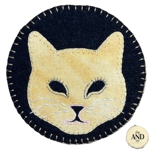 May include: A round, black felt coaster with a stitched border. The center features a light tan cat face with black eyes, nose, and stitched whiskers. The coaster has a small, circular logo in the bottom right corner.