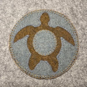 Könnte beinhalten: Eine Filzapplikation einer braunen Schildkröte auf blauem Hintergrund. Die Schildkröte ist mit goldfarbenem Garn umrandet.