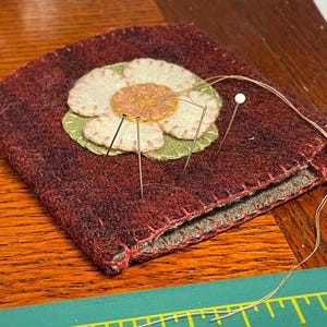 May include: A red felt pincushion with a green and white flower design. The pincushion is filled with pins and a white bead.