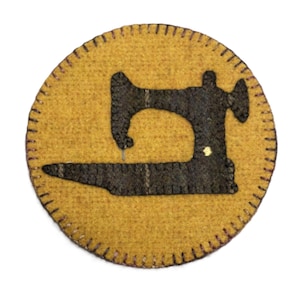 May include: A round, mustard-yellow felt patch with a black sewing machine silhouette in the center. The patch has a stitched border with purple thread. A small gold-colored detail is visible on the sewing machine.