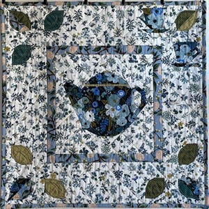 May include: A square quilt featuring a central teapot design surrounded by floral patterns in shades of blue, white, and green. The quilt has a quilted texture and a light blue border, with appliqued leaves and teacups in the corners.