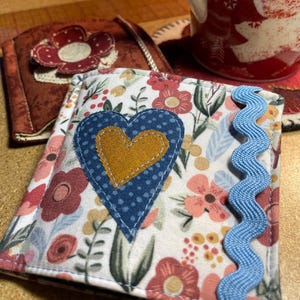 Puede incluir: Un calientavasos de tela blanca con un corazón azul y un centro amarillo. El corazón tiene lunares blancos. El calientavasos tiene un ribete azul festoneado.