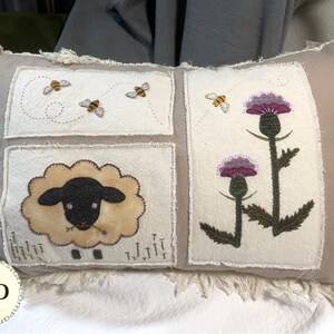 Peut inclure: Un coussin rectangulaire beige avec des bords effilochés. Il présente des panneaux brodés avec des abeilles, un mouton et des chardons violets. Le design a une esthétique rustique et faite à la main, avec des éléments naturels.