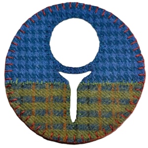May include: A circular, handmade textile item with a large central opening and a smaller, downward-pointing extension. The top half is blue with a textured pattern, while the bottom half is green and orange plaid. Red stitching outlines the edges.