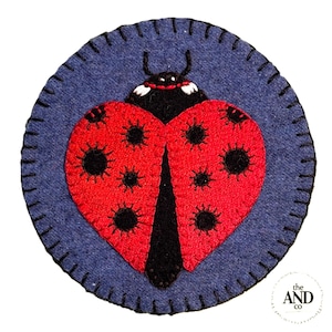May include: A round, blue felt patch with a red ladybug design. The ladybug has black spots and a black head. The patch is stitched with black thread around the edge. The logo "the AND co" is in the bottom right corner.