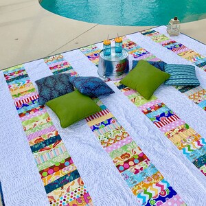 May include: A white and blue patterned picnic blanket with colorful stripes, laid out on a concrete patio next to a swimming pool. There are four pillows on the blanket, two green, one blue, and one dark blue. There is a metal bucket with two glass jars on top of it.