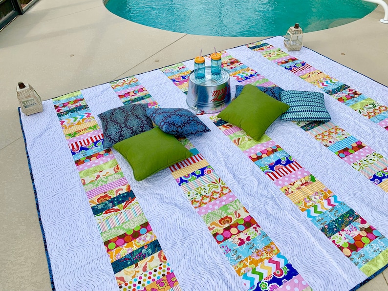 May include: A white and blue patterned picnic blanket with colorful stripes, laid out on a concrete patio next to a swimming pool. There are four pillows on the blanket, two green, one blue, and one dark blue. There is a metal bucket with two glass jars on top of it.