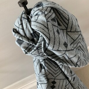 May include: A gray scarf with a geometric black pattern is draped over a black metal hook. The scarf is knotted, showcasing the intricate design. The fabric appears soft and textured, with a subtle heathered effect.