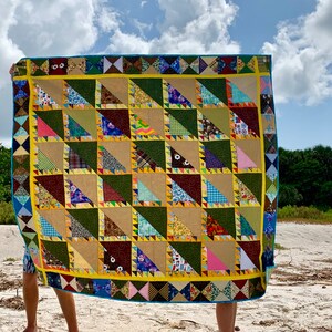 May include: A colorful patchwork quilt with a geometric pattern of triangles. The quilt is made of various fabrics in shades of green, brown, yellow, pink, blue, and white. The quilt is being held up by a person on a sandy beach.