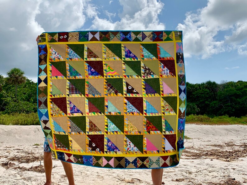 May include: A colorful patchwork quilt with a geometric pattern of triangles. The quilt is made of various fabrics in shades of green, brown, yellow, pink, blue, and white. The quilt is being held up by a person on a sandy beach.