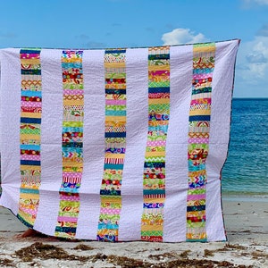 May include: A colorful patchwork quilt with white stripes and a variety of colorful fabrics. The quilt is made up of strips of fabric sewn together to create a striped pattern. The quilt is displayed on a beach with the ocean in the background.