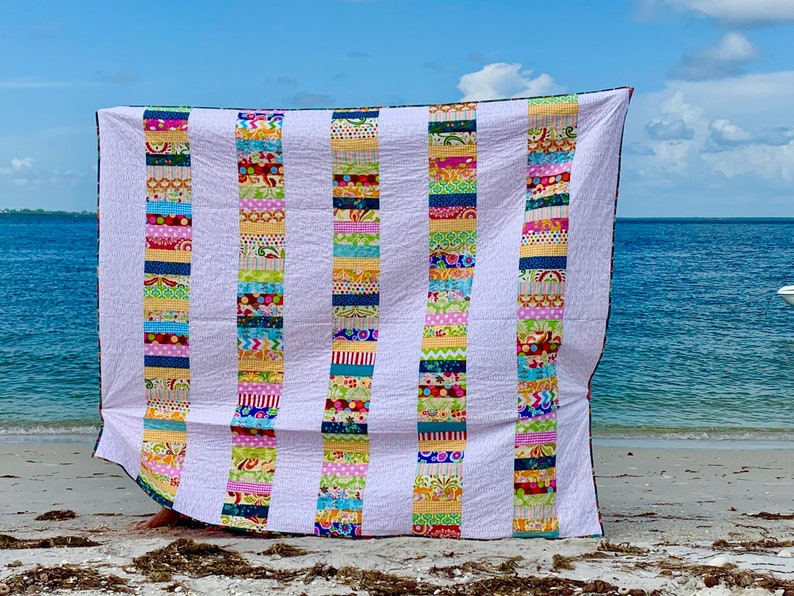 May include: A colorful patchwork quilt with white stripes and a variety of colorful fabrics. The quilt is made up of strips of fabric sewn together to create a striped pattern. The quilt is displayed on a beach with the ocean in the background.