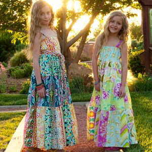 K&ouml;nnte beinhalten: Zwei junge M&auml;dchen tragen bunte, gemusterte Maxikleider. Die Kleider haben ein Patchwork-Design mit verschiedenen floralen und geometrischen Mustern. Die M&auml;dchen stehen auf einem Weg mit gr&uuml;nem Gras und B&auml;umen im Hintergrund.