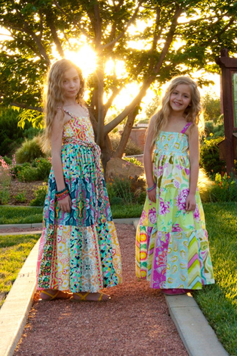 K&ouml;nnte beinhalten: Zwei junge M&auml;dchen tragen bunte, gemusterte Maxikleider. Die Kleider haben ein Patchwork-Design mit verschiedenen floralen und geometrischen Mustern. Die M&auml;dchen stehen auf einem Weg mit gr&uuml;nem Gras und B&auml;umen im Hintergrund.