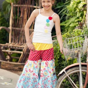 May include: A young girl wearing a white tank top with colorful flower embellishments and a multi-colored, tiered skirt. The skirt has a red, yellow, and blue pattern with white polka dots. The girl is standing next to a bicycle with a basket.