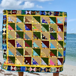 May include: A colorful patchwork quilt made of various fabrics and patterns, featuring a mix of triangles and squares. The quilt is held up in front of a person standing on a sandy beach.