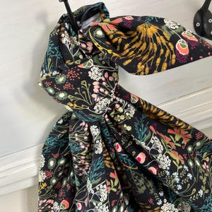 May include: A black fabric head wrap with a colorful floral pattern, including white, pink, yellow, and green accents. The head wrap is tied in a bow and hangs from a black hook on a white painted surface.