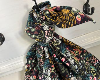 May include: A black fabric head wrap with a colorful floral pattern, including white, pink, yellow, and green accents. The head wrap is tied in a bow and hangs from a black hook on a white painted surface.