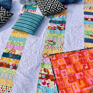 May include: A white quilt with a patchwork design featuring colorful stripes and floral patterns. The quilt is folded in half and has a pink and orange geometric pattern on the back.