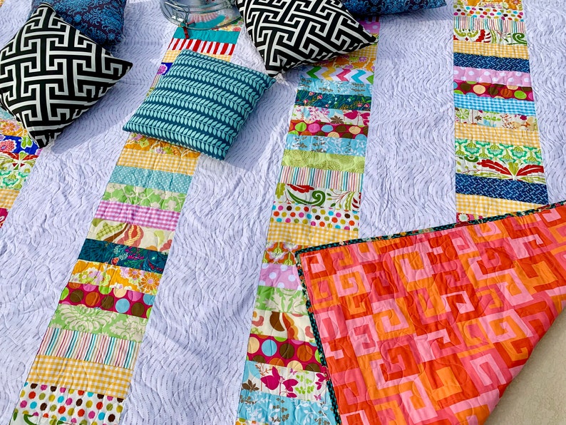 May include: A white quilt with a patchwork design featuring colorful stripes and floral patterns. The quilt is folded in half and has a pink and orange geometric pattern on the back.