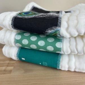 May include: Close-up of three white, textured baby washcloths. Each cloth has a decorative band with a different pattern: a peacock feather design, white polka dots on green, and a teal pattern. The bands are stitched with white thread.