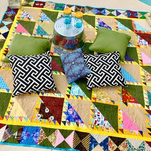May include: A colorful patchwork quilt with a geometric pattern of triangles. The quilt is spread out on the ground and has several pillows on top of it. There are two glass bottles and a metal bucket on the quilt.
