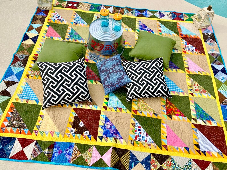 May include: A colorful patchwork quilt with a geometric pattern of triangles. The quilt is spread out on the ground and has several pillows on top of it. There are two glass bottles and a metal bucket on the quilt.