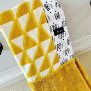 Op de afbeelding: Een gele badjas met een witte en zwarte geometrische print. De badjas heeft een wit label met de tekst "Alhambra" erop.