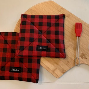 Puede incluir: Dos agarraderas a cuadros rojos y negros con un diseño cosido. Un pincel de silicona rojo y una pala de pizza de madera también están en la imagen. Las agarraderas tienen una pequeña etiqueta negra con texto blanco.