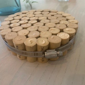 May include: A round coaster made from a collection of wine corks, secured together with a metal clamp. The corks are arranged in a circular pattern, creating a unique and rustic design.