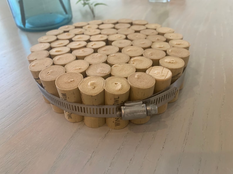 May include: A round coaster made from a collection of wine corks, secured together with a metal clamp. The corks are arranged in a circular pattern, creating a unique and rustic design.