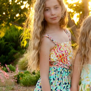 K&ouml;nnte beinhalten: Ein junges M&auml;dchen mit langen, welligen blonden Haaren tr&auml;gt ein farbenfrohes, gemustertes Sommerkleid mit einem leuchtend pinken, gelben und blauen Design. Das Kleid hat einen R&uuml;schen-Saum und eine Schleife an der Taille.