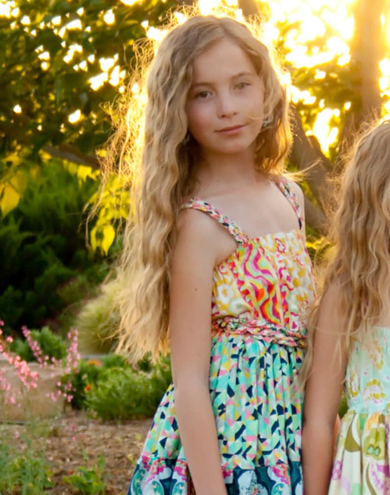 K&ouml;nnte beinhalten: Ein junges M&auml;dchen mit langen, welligen blonden Haaren tr&auml;gt ein farbenfrohes, gemustertes Sommerkleid mit einem leuchtend pinken, gelben und blauen Design. Das Kleid hat einen R&uuml;schen-Saum und eine Schleife an der Taille.