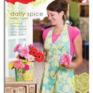 May include: A teal and white floral print halter apron with the text "daily spice Heather Bailey Living Patterns" on the front. The apron is being worn by a woman with brown hair and a pink shirt.