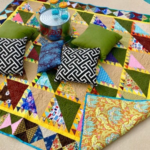 May include: A patchwork quilt with a variety of colors and patterns, including green, brown, yellow, blue, and pink. The quilt is made of squares and triangles, and it has a scalloped edge. There are four pillows on the quilt, two green, one black and white, and one blue. A metal bucket with the word "Coca-Cola" on it is in the background.