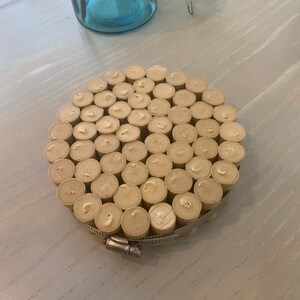 May include: A round coaster made from a collection of wine corks, secured with a metal clamp. The coaster is light brown and has a natural, rustic look.