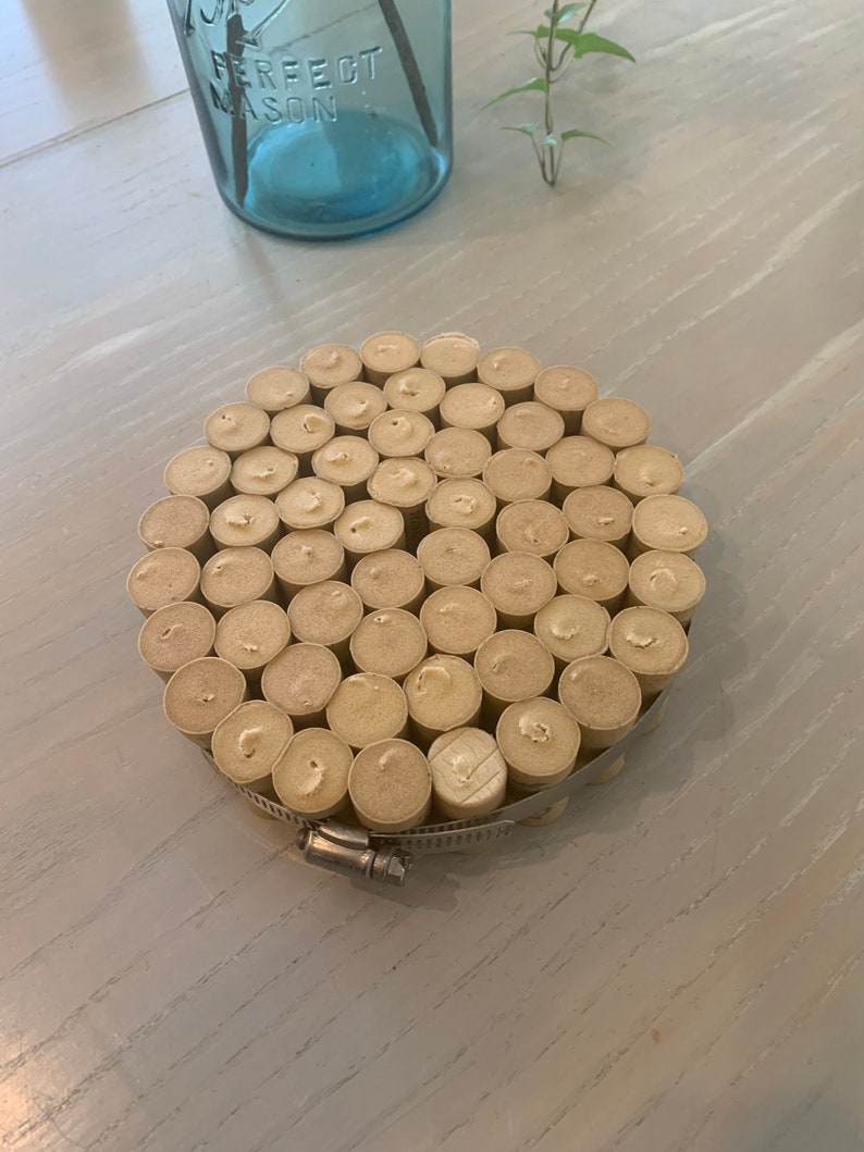 May include: A round coaster made from a collection of wine corks, secured with a metal clamp. The coaster is light brown and has a natural, rustic look.