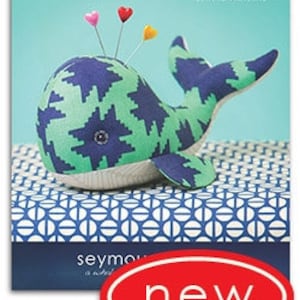 Puede incluir: Un juguete de peluche de ballena azul turquesa y azul marino con un patrón geométrico. La ballena está sentada en una caja con estampado azul y blanco con el texto "seymour is a whale" y la palabra "new" en un óvalo rojo.