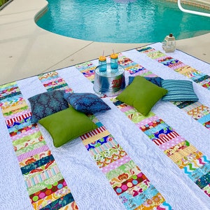 May include: A white and blue patchwork picnic blanket with colorful stripes and patterns. The blanket is laid out on a patio next to a swimming pool. There are four pillows on the blanket, two green, one dark blue, and one purple. There are two glass jars with yellow lids and straws on a metal bucket.