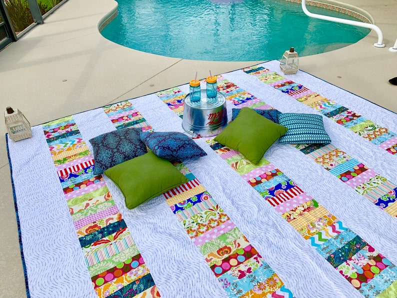 May include: A white and blue patchwork picnic blanket with colorful stripes and patterns. The blanket is laid out on a patio next to a swimming pool. There are four pillows on the blanket, two green, one dark blue, and one purple. There are two glass jars with yellow lids and straws on a metal bucket.