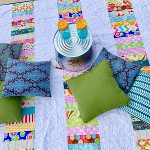 May include: A white patchwork quilt with colorful stripes and patterns. There are five blue and gray patterned throw pillows and one green throw pillow on the quilt. A metal container with two glass jars with orange lids and straws is in the center of the quilt.