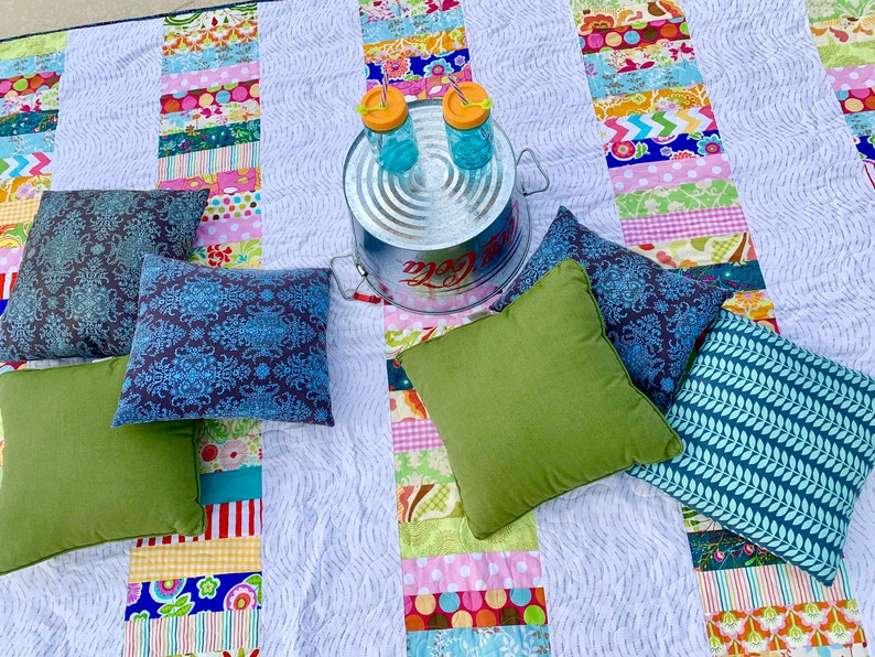 May include: A white patchwork quilt with colorful stripes and patterns. There are five blue and gray patterned throw pillows and one green throw pillow on the quilt. A metal container with two glass jars with orange lids and straws is in the center of the quilt.